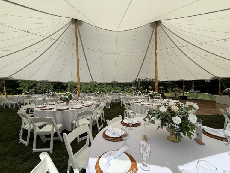 Round tables with white linens, woven chargers and floral centerpieces inside a sailcloth tent